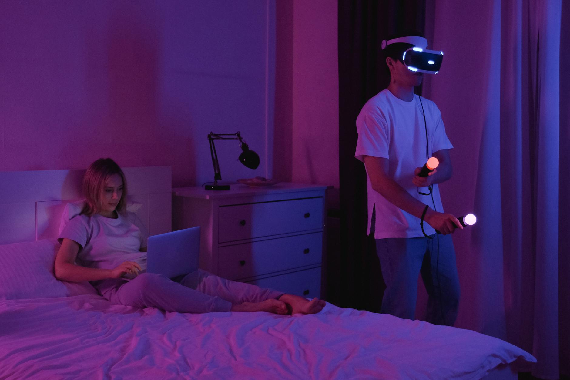 Mężczyzna w goglach VR i kobieta z laptopem w przyciemnionej sypialni